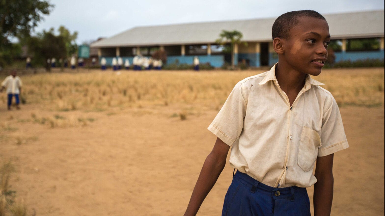 Young boy in school uniform looks off to the side with school in background