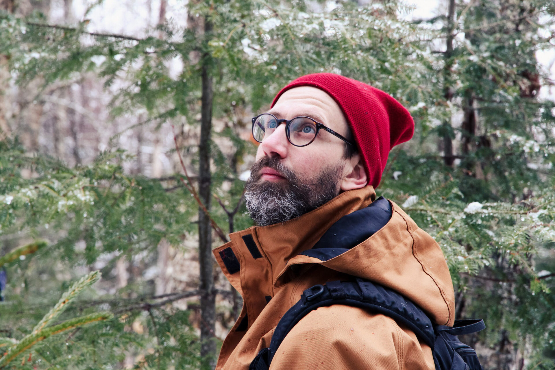 A man with round glasses and a red hat looks up at the forest canopy.