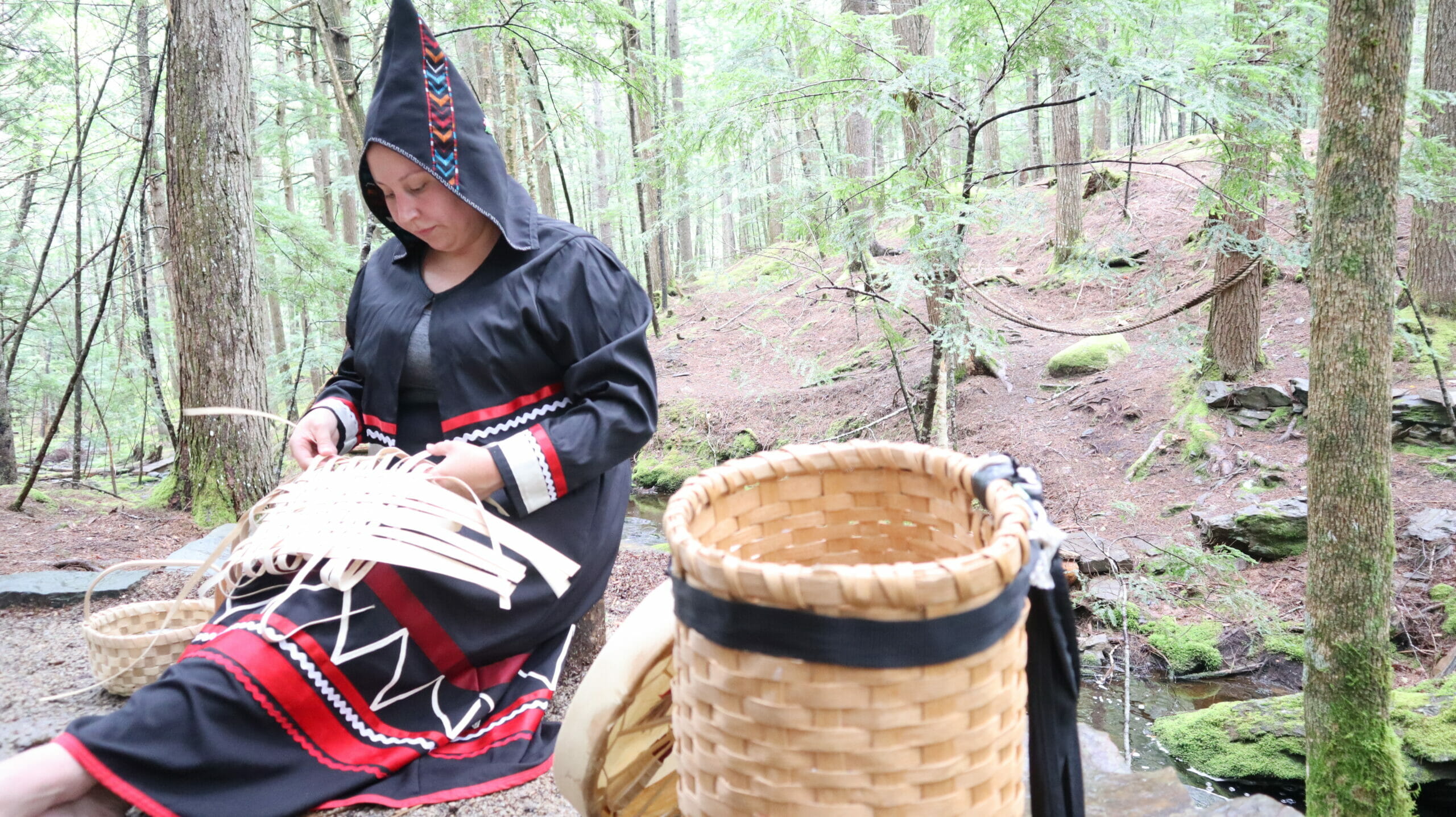 Photo of a woman dressed in black and red traditional Mi'kmaq regelia as she sits in a forest clearing weaving a basket from pale yellow ash bark. 