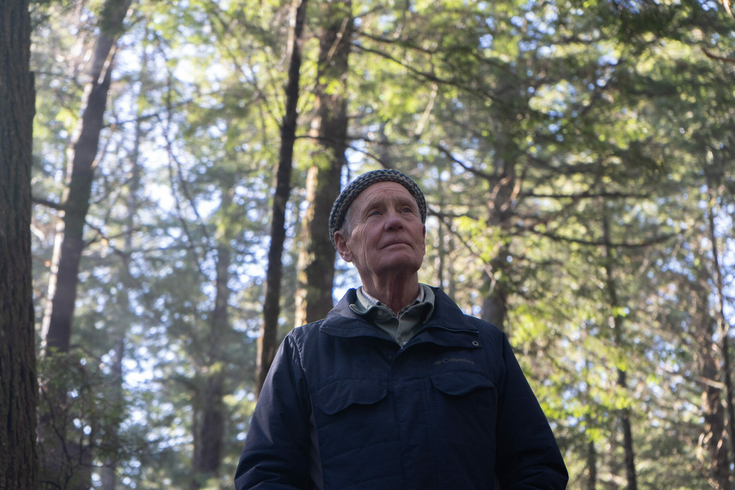 An older man wearing a woolen hat and blue jacket stands in with his hands in his pockets, looking past the camera into the distance. Behind him, sunlight filters through a green forest canopy.